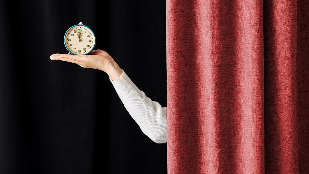 A hand holding an alarm clock appearing from behind a red curtain.