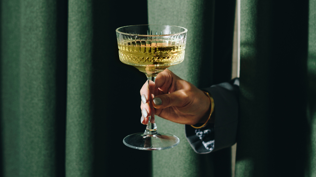 A hand holding a cocktail appearing between green curtains.